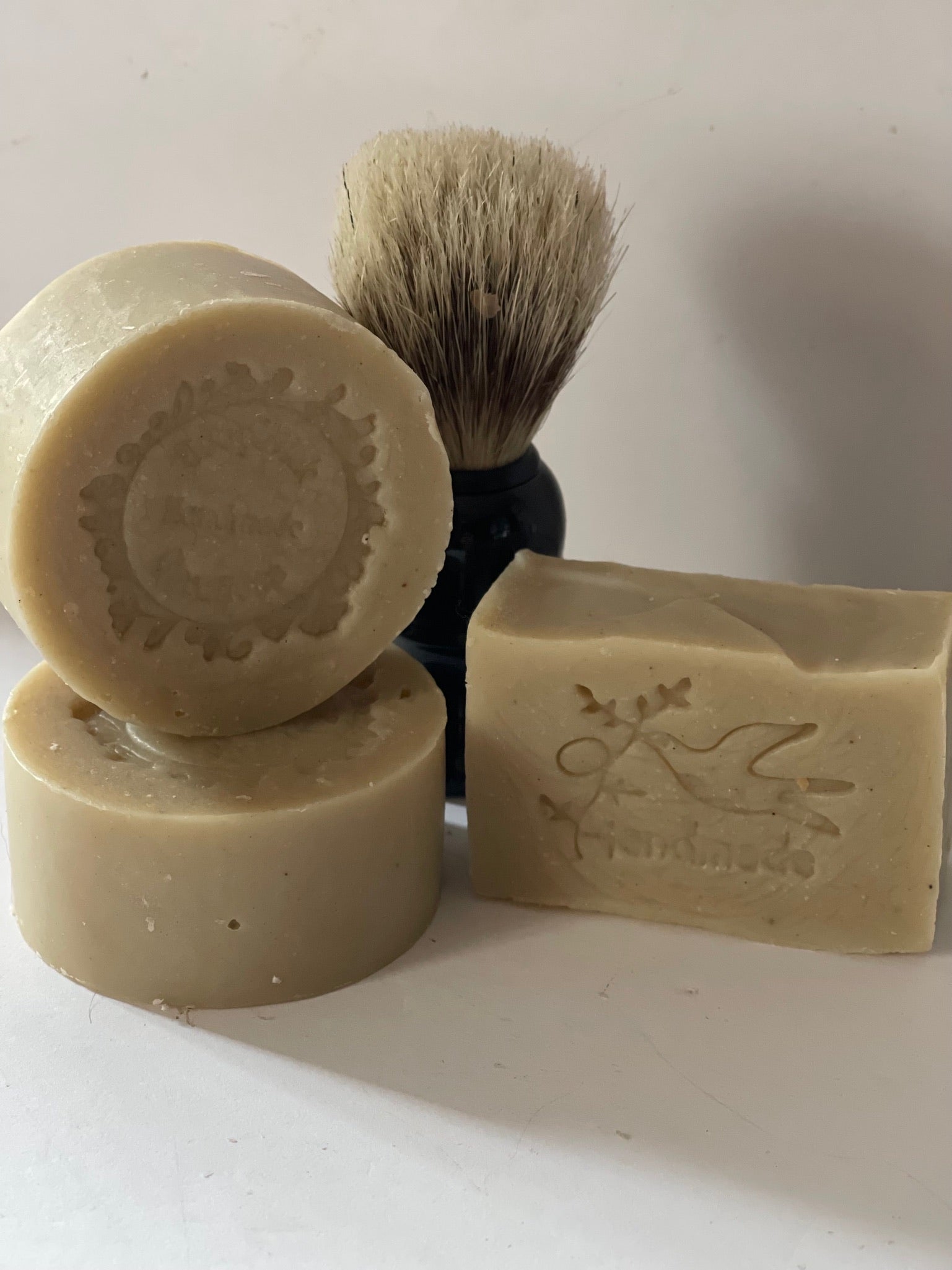 Photograph of a beige  two round men’s soap pucks and bar soap pictured with a shaving brush in the background