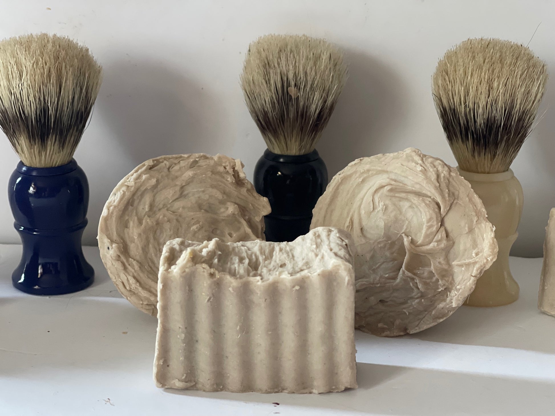 Photo of dual lye, tobacco and rum, shaving bars, and pucks with shaving brushes in the background.